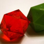 Photograph of two twenty sided dice from GameScience, one opaque green and one translucent red. The 7 face in on top of both, and flashing is visible along one edge of the 7 face.